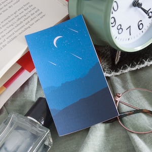 Puede incluir: Una tarjeta azul con una luna creciente y estrellas fugaces sobre un fondo de montañas azules oscuras. La tarjeta está sobre una superficie de tela verde con un libro, un reloj despertador y una botella de perfume.