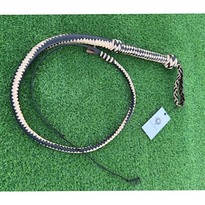 May include: A black and tan leather bullwhip with a braided handle. The whip is coiled on a green surface.