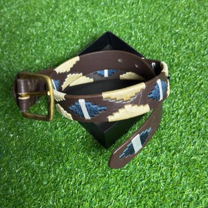 May include: A brown leather belt with a beige and blue embroidered pattern. The belt has a gold buckle.