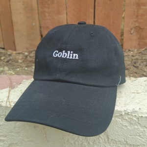 May include: Black baseball cap with the word "Goblin" embroidered in white. The cap has a curved brim and a button on top. The background features a wooden fence and a textured, off-white surface.