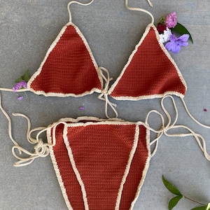 May include: A rust-colored crochet bikini with a white trim. The bikini top is a triangle style with adjustable straps. The bikini bottom is a high-cut style with ties on the sides.