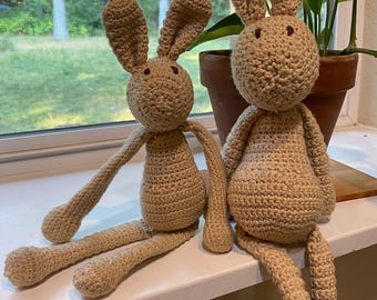 May include: Two handmade crochet rabbits in a light tan yarn. Each rabbit features long, upright ears, a round head with dark brown embroidered eyes, a slender torso, and long, dangling arms and legs. The rabbits are crafted with a visible stitch pattern.