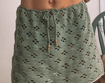 May include: A sage green crochet skirt with a floral pattern and a drawstring waist. The skirt has a scalloped edge and is made of a soft, textured yarn. The skirt is a mini length.