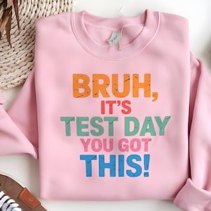 May include: Pink sweatshirt with the text "Bruh, it's test day you got this!" in colorful letters.