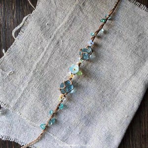 May include: A bracelet featuring a brown cord base adorned with two large, faceted light blue stones, smaller light blue and green beads, and tiny pale blue flower-shaped accents. The design incorporates a mix of textures and colours.