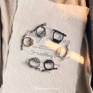 May include: A collection of handmade rings with various gemstones, including quartz and amethyst, set in knotted cord. The rings are arranged on a textured paper with wedding-themed text. The rings are in shades of brown, black, and gray.