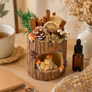 Puede incluir: Un recipiente cilíndrico con forma de tocón de árbol que presenta un centro hueco con figuras de osos en miniatura y setas naranjas. La parte superior está decorada con ramas de canela, rodajas de limón seco, una piña, bellotas y follaje verde.