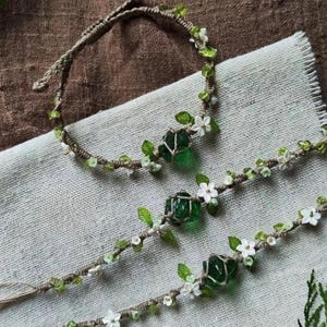 May include: A handcrafted jewellery set comprising a headband and two bracelets. Each piece is decorated with green glass stones, small white flowers, and green leaves, set on a natural twine base. The text "Herbarumart Studio" is visible.