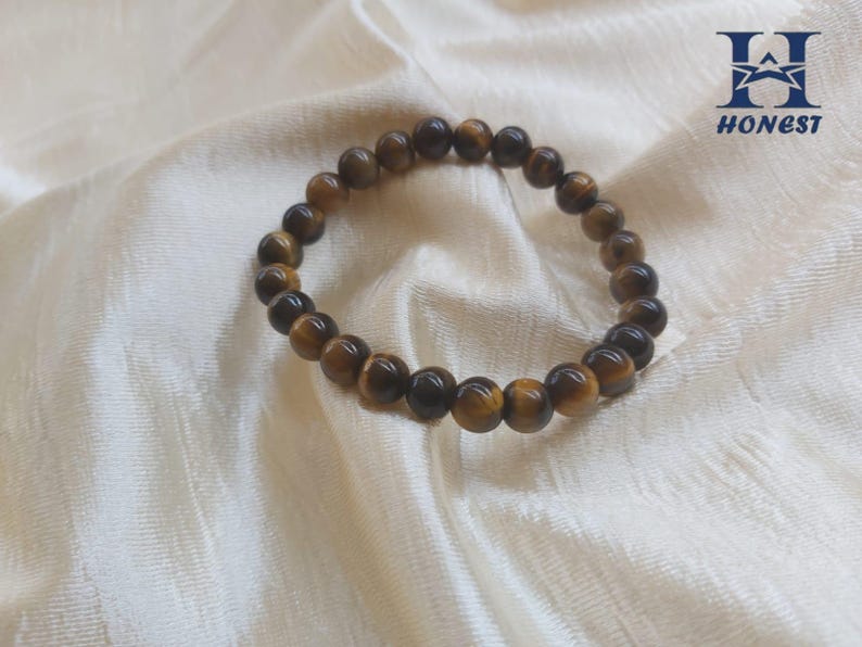 May include: A beaded bracelet made of tiger's eye gemstones. The bracelet features round, polished beads in shades of brown and gold. The bracelet is displayed on a white, textured fabric.