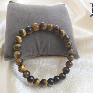 May include: A beaded bracelet made of tiger's eye gemstones. The bracelet features a mix of dark brown and golden-brown beads, strung together to form a circular shape. The bracelet is displayed on a gray pillow and a white fabric background.