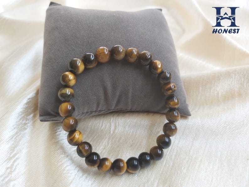 May include: A beaded bracelet made of tiger's eye gemstones. The bracelet features a mix of dark brown and golden-brown beads, strung together to form a circular shape. The bracelet is displayed on a gray pillow and a white fabric background.