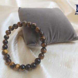 May include: A beaded bracelet made of tiger's eye gemstones. The bracelet is a circular shape, with each bead a warm brown color with golden and black streaks. The bracelet is resting on a white surface with a gray pillow in the background.