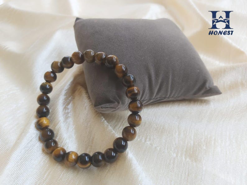 May include: A beaded bracelet made of tiger's eye gemstones. The bracelet is a circular shape, with each bead a warm brown color with golden and black streaks. The bracelet is resting on a white surface with a gray pillow in the background.