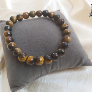 May include: A beaded bracelet made of tiger's eye gemstones. The bracelet features round, polished beads in shades of brown and gold. The bracelet is resting on a gray pillow. The word "HONEST" is printed on the upper right corner.