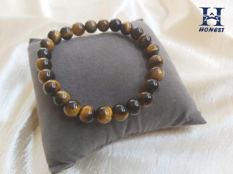 May include: A beaded bracelet made of tiger's eye gemstones. The bracelet features round, polished beads in shades of brown and gold. The bracelet is resting on a gray pillow. The word "HONEST" is printed on the upper right corner.