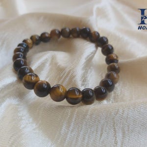 May include: A beaded bracelet featuring round, polished tiger's eye stones in shades of brown and gold. The bracelet is strung on an elastic band and rests on a cream-colored, textured fabric.