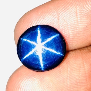 Op de afbeelding: Een ronde, blauwe ster-saffier edelsteen met een zespuntige ster in het midden. De ster is wit en de saffier is diepblauw.
