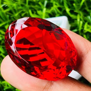 Peut inclure: Une pierre précieuse rouge vif, taillée en forme ovale. La gemme est tenue dans une main sur un fond d'herbe verte. La taille de la pierre crée de multiples surfaces réfléchissantes, rehaussant son éclat. Un exemple frappant de joaillerie.