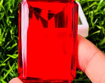 May include: A large, deep red, faceted gemstone with a rectangular shape. The gemstone is held in a person's hand.