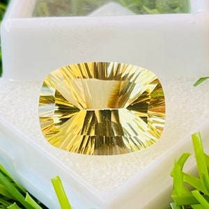 Puede incluir: Una gran piedra preciosa de citrino de forma ovalada, facetada, de color dorado, con un aspecto brillante, claro y translúcido. La piedra preciosa está colocada en una caja blanca sobre un fondo blanco.