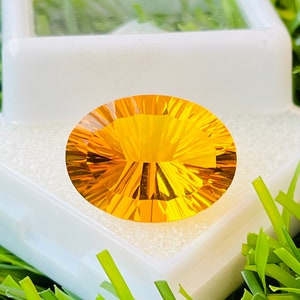 Puede incluir: Una gema facetada, de corte ovalado, con un vibrante tono amarillo-naranja. La gema se exhibe en una caja de plástico blanca, sobre un fondo de césped artificial verde. Los intrincados cortes de la gema crean un efecto radiante.