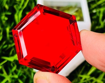 May include: A bright red hexagonal gemstone with a faceted cut. The gemstone is held in a person's hand.