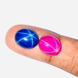 May include: Two oval-shaped gemstones, one blue and one pink, with a star-like pattern inside. The stones are held by a finger.