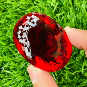 May include: A large, faceted, red gemstone with a high polish. The gemstone is oval shaped and has a checkerboard pattern on the surface.