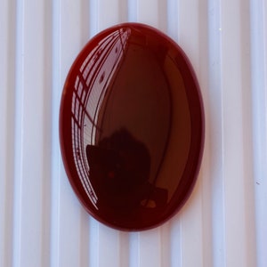 Puede incluir: Una piedra de cornalina roja redonda y lisa con un alto pulido. La piedra tiene forma ovalada y un color rojo intenso.
