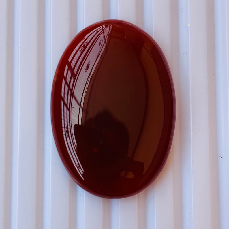 Puede incluir: Una piedra de cornalina roja redonda y lisa con un alto pulido. La piedra tiene forma ovalada y un color rojo intenso.