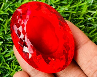 May include: A large, oval-shaped, red gemstone with a faceted cut. The gemstone is held in a hand, with a green grassy background.