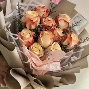 May include: A bouquet of peach and coral roses, accented with greenery, wrapped in layers of brown and pink paper. The roses are in full bloom, showcasing their delicate petals. A beautiful floral arrangement.