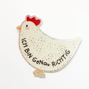 Può includere: Un pollo bianco con una cresta rossa e dettagli glitterati. Il pollo tiene un cartello che dice "Ich Bin Genau Richtig" in testo nero.