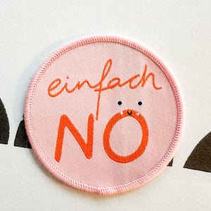 Könnte beinhalten: Ein runder, rosa Aufnäher mit den deutschen Worten "einfach NO" in orange gestickt. Das "O" in "NO" hat Cartoon-Augen und einen Mund. Der Aufnäher hat einen gestickten Rand.