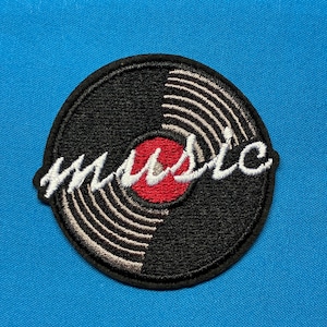 May include: A circular embroidered patch featuring a black vinyl record design with concentric silver-toned grooves and a solid red centre. The word music is stitched across the centre in a white, cursive, lowercase script font.