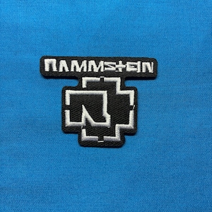 Puede incluir: Parche bordado con el nombre de la banda "RAMMSTEIN" sobre un símbolo de cruz estilizado. El parche tiene un borde negro con costuras blancas para el texto y el símbolo, sobre un fondo azul brillante.