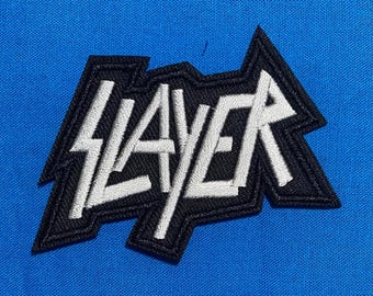 以下が含まれることがあります： 黒い縁取りが施された白い糸で「SLAYER」という文字が刺繍されたワッペン。文字は鋭く角張ったエッジと独特のフォントデザインで構成されています。ワッペンは無地の鮮やかな青い布地の上に配置されています。