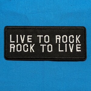 May include: A rectangular black embroidered patch with a stitched border. The text "LIVE TO ROCK" is embroidered in white capital letters on the top line, and "ROCK TO LIVE" is embroidered in white capital letters on the bottom line.