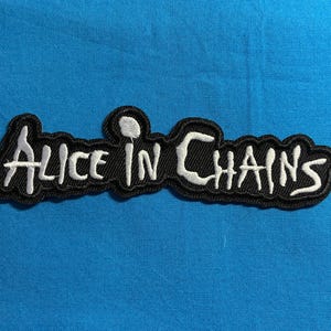 May include: A black embroidered patch featuring the white text Alice in Chains in a stylised font. The patch has a thick black border that follows the contour of the lettering, creating a distinct outline against the background fabric.