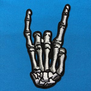 Könnte beinhalten: Ein gestickter Aufnäher mit einer weißen und grauen Skeletthand, die ein Rock-and-Roll-Zeichen mit ausgestrecktem Zeige- und kleinen Finger zeigt. Die detaillierte Skeletthand ist schwarz umrandet und auf einem einfarbigen, hellblauen Stoff.