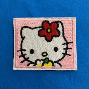 May include: An iron-on patch featuring Hello Kitty with a red flower on her head and a white flower in her paw, embroidered in pink and white.