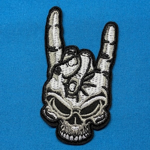 Könnte beinhalten: Ein gestickter Aufnäher, der einen Totenkopf mit einer aus der Oberseite ragenden Hand zeigt, die eine Rock-and-Roll-Geste formt. Das Design verwendet weißen, grauen und schwarzen Faden für Tiefe und Schattierung, mit einem dicken schwarzen Rand.