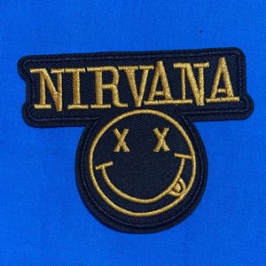 May include: A black embroidered patch featuring the word NIRVANA in gold thread above a circular smiley face icon with X-shaped eyes and a tongue sticking out, all set against a vibrant, solid blue fabric background.