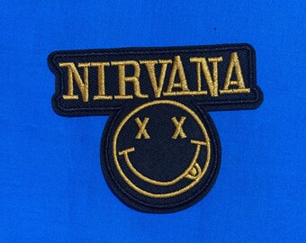 May include: A black embroidered patch featuring the word NIRVANA in gold thread above a circular smiley face icon with X-shaped eyes and a tongue sticking out, all set against a vibrant, solid blue fabric background.