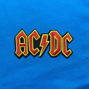 May include: An embroidered patch featuring the AC/DC logo in red with a yellow border on a bright blue fabric background. The design includes the letters A, C, D, and C with a central lightning bolt symbol, all outlined in black thread.