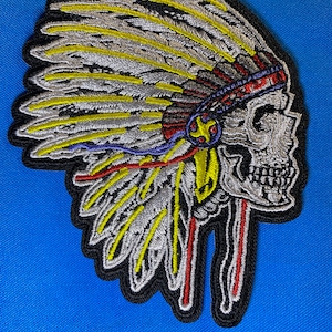 May include: A black and white embroidered patch of a skull wearing a headdress with yellow, red, and blue feathers.