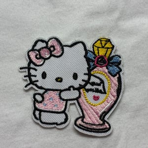 Pode incluir: Patch bordado com um gato de desenho animado com laço e vestido rosa, segurando um frasco de perfume rosa com uma gema amarela. O patch tem contornos pretos em um fundo branco.