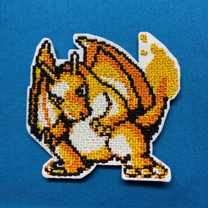 May include: An embroidered patch featuring a pixelated, orange and yellow dragon-like creature with a flame on its tail. The design uses black outlines and white highlights to define the shape of the creature against a solid blue background.