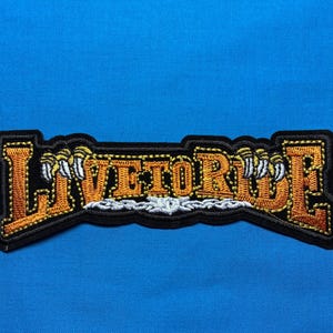 Può includere: Toppa ricamata con le parole "LIVE TO RIDE" in filo arancione e bianco, bordata di nero. La toppa presenta artigli e un bordo nero, su sfondo blu.