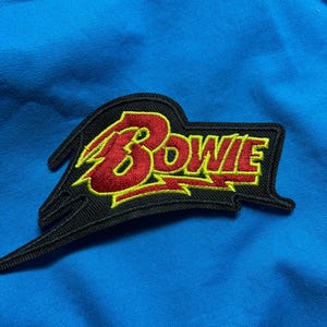 May include: An embroidered patch featuring the word BOWIE in red lettering with a yellow outline. The text is set against a black background shaped like a lightning bolt, with a sharp, angular design that extends to the left and right sides.
